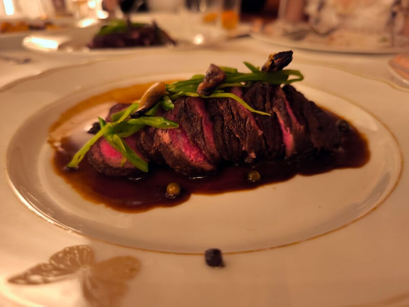 A beautifully plated dish featuring sliced, medium-rare steak topped with green vegetables and mushrooms, served in a rich, dark sauce on an elegant white plate with a butterfly design. The background shows a softly lit dining setting.