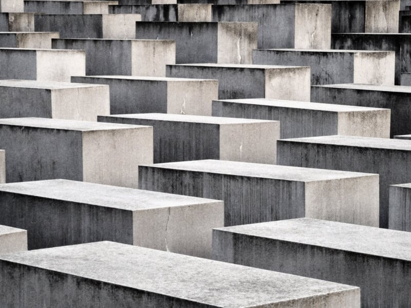 The image shows a large array of grey concrete slabs of varying heights. They are arranged in a grid pattern, creating a sense of depth and perspective. The slabs are part of a memorial with a stark, solemn appearance.