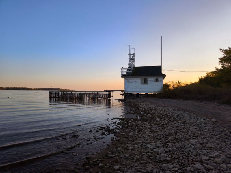 A tranquil lakeside scene at sunset shows a small white boathouse on stilts, with a ladder and antenna, next to a rocky shore. The calm water reflects the clear evening sky, transitioning from light blue to warm hues near the horizon.