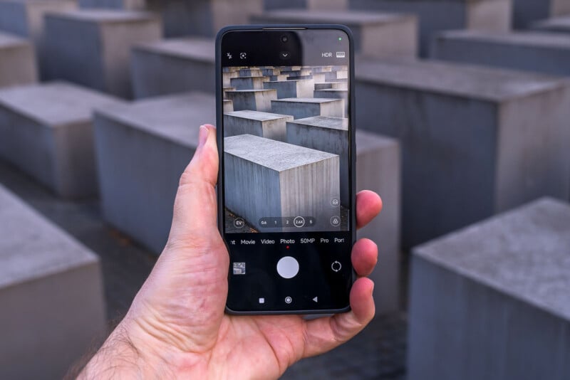 A hand holds a smartphone displaying a camera app interface. The phone's screen shows a live view of a grid of large, concrete blocks, similar to the blocks surrounding the person holding the phone.