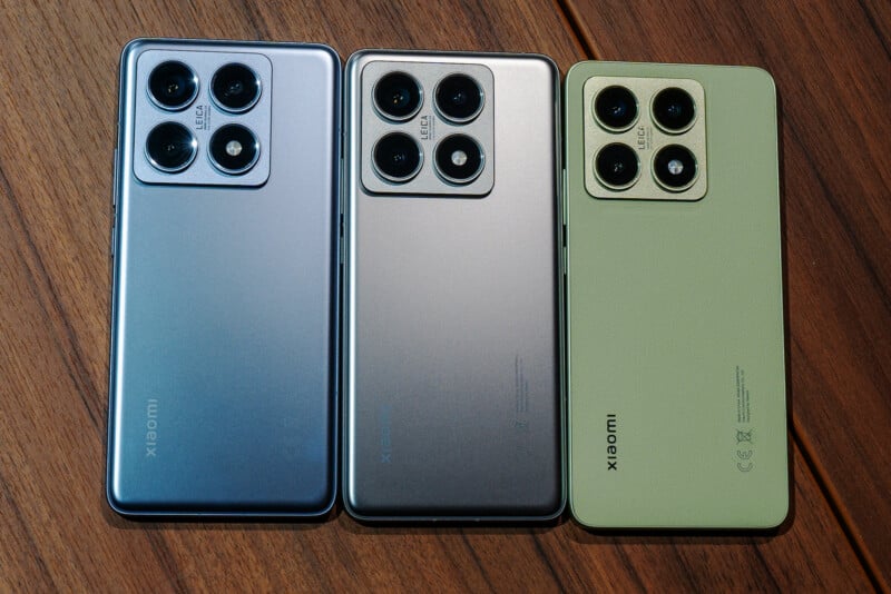 Three smartphones in different colors (blue, gray, and green) are displayed side by side on a wooden surface. Each phone features a rectangular camera block with three lenses and an LED flash in the top left corner.