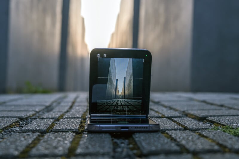 A foldable smartphone is placed on a cobblestone pathway, capturing a photo of a long, narrow alleyway bordered by tall concrete walls. The screen displays the perspective of the alley, emphasizing the symmetry and depth of the scene.