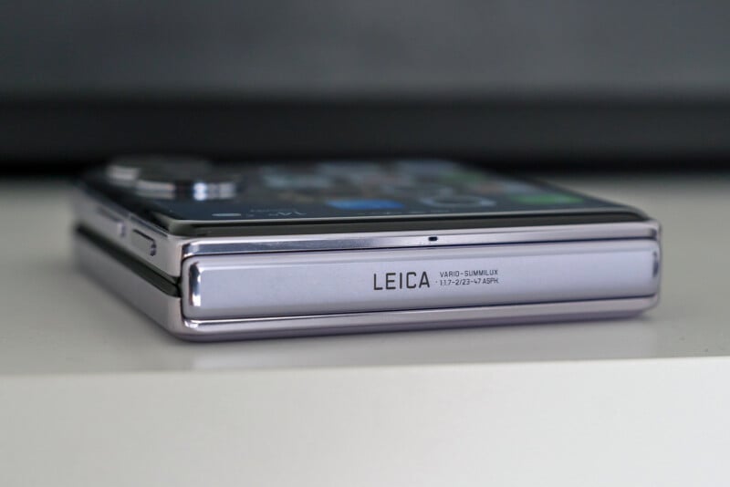 Close-up of a folded smartphone with a reflective surface, resting on a white surface. The brand name "Leica" and the camera specifications "Vario-Summilux 1:1.9-2.4/19-57 ASPH." are visible on the side of the device.