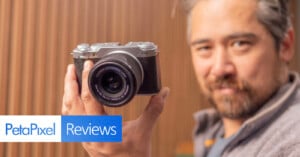 A person holding a vintage-style camera close to the lens, standing against a blurred wooden background. The image features the text "PetaPixel Reviews" in the bottom left corner.