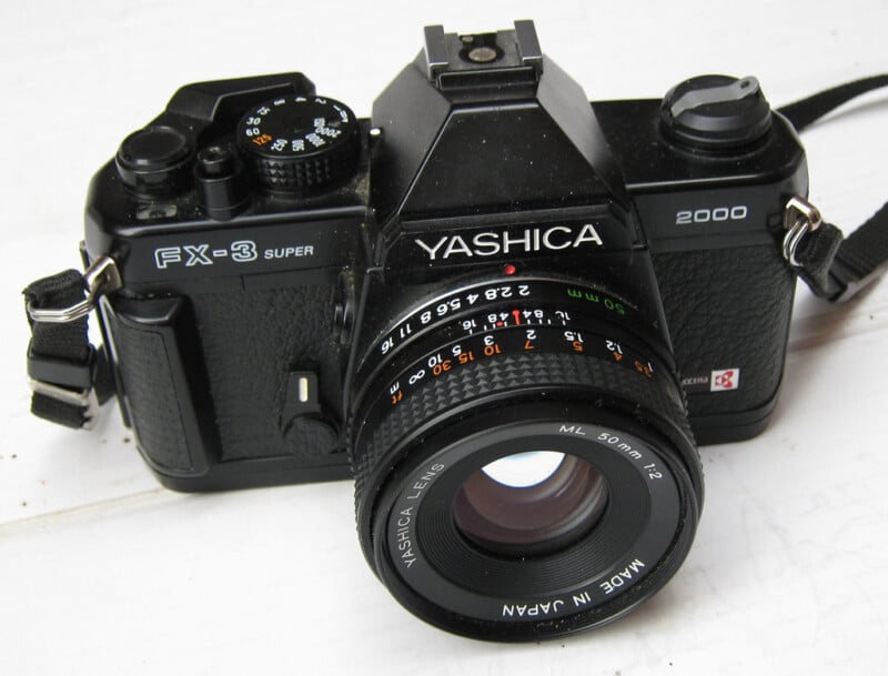 A black Yashica FX-3 Super 2000 film camera with a 50mm Yashica lens is placed on a white surface. The camera features a textured grip, visible dials, and a strap attached on each side.