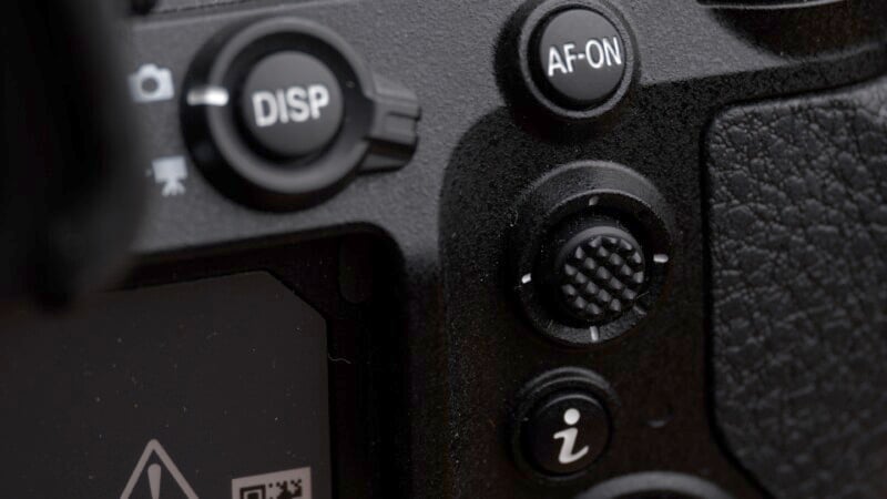 Close-up of a black digital camera's control buttons. Visible buttons include "DISP," "AF-ON," a textured joystick, and an "info" button labeled with an "i." The edge of the camera's screen is also partially visible. The camera's body has a textured grip.