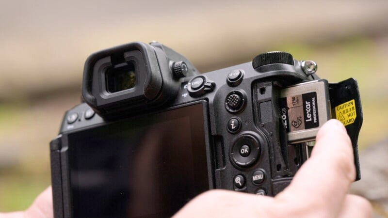 A close-up image of a person inserting a memory card into a DSLR camera. The camera's LCD screen and various control buttons are visible. The memory card has a "Caution: Hot" label on it. Blurry background.