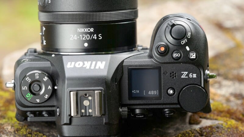 Close-up image of a Nikon Z6 II camera with a 24-120mm f/4 S lens. The camera is resting on a mossy rock. The view shows the top part of the camera including buttons and controls, along with a small screen displaying settings.