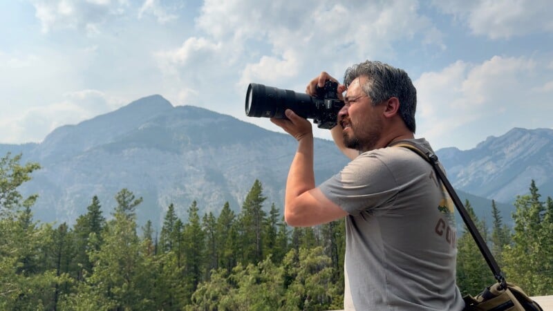 A man with short gray hair and glasses is photographing with a DSLR camera on a sunny day. He stands in front of a vast mountain range covered in greenery, with a forest in the foreground and a partly cloudy sky in the background.