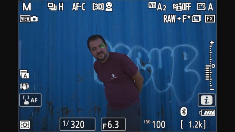 A man wearing a maroon t-shirt stands in front of a blue wall with graffiti, framed through a camera's viewfinder. The camera settings are visible, including ISO 100, aperture f/6.3, shutter speed 1/320, and a green focus square on the man's eye.