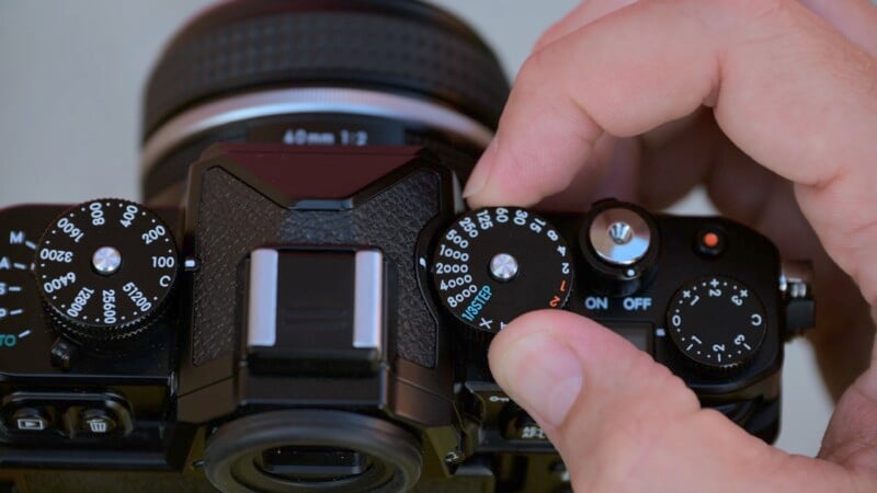 A close-up view of a person adjusting the settings on a vintage-style camera. The focus is on the top dials, including ISO sensitivity, shutter speed, and other controls, showcasing the intricate details of the camera's interface and the person's fingers turning a dial.