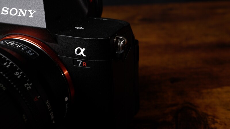 Close-up of a Sony Alpha 7R camera, showing the brand logo and red "7R" label. The camera is resting on a wooden surface, partially in shadow, highlighting its sleek, black design and lens details.