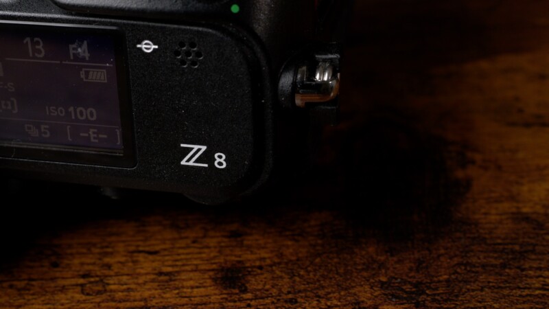 Close-up of a black camera body with "Z 8" printed on it, partially showing a screen with camera settings. The surface under the camera is a textured wooden surface.