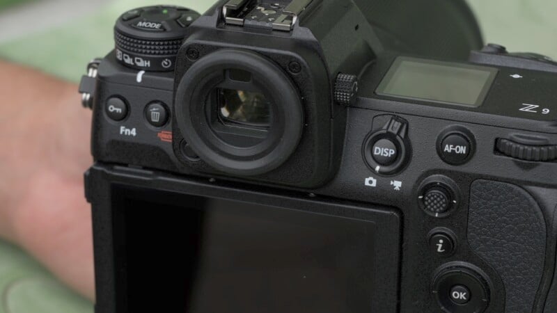 Close-up of a black digital camera showing buttons and dials on the back, including the viewfinder, DISP, and AF-ON buttons. The camera is positioned on a green surface.