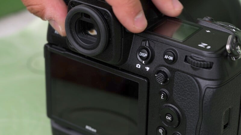 Close-up of a person holding a black camera, adjusting buttons near the viewfinder. The camera's display screen and various control buttons are visible.