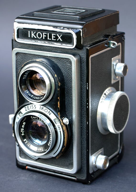 A vintage Ikoflex twin-lens reflex camera is shown against a plain background, featuring two lenses, a focus knob, and a rectangular viewfinder on the top. The camera has a classic black and silver design.