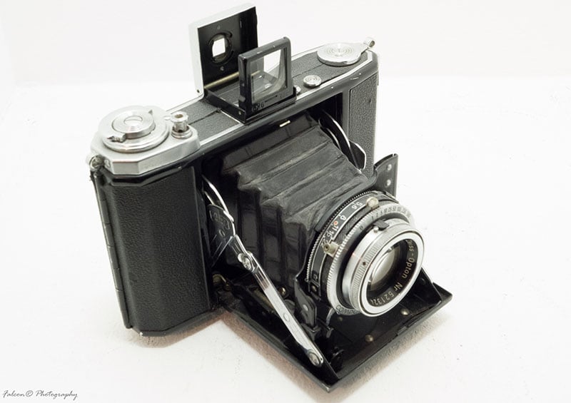 A vintage folding camera with a bellows design, featuring a black body and silver dials. The lens is extended outward, and a metal frame viewfinder is visible on top. The camera rests on a white background.