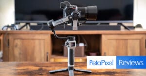 A professional camera is mounted on a stabilizing gimbal, which is placed on a wooden surface. In the background is a wooden console with a television. The photo has overlapping text that reads "PetaPixel Reviews" in the lower right corner.