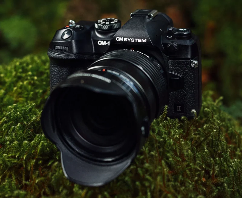 A black OM System camera with a large lens rests on a lush green mossy surface. The detailed texture of the moss contrasts with the sleek design of the camera, highlighting its features and branding.