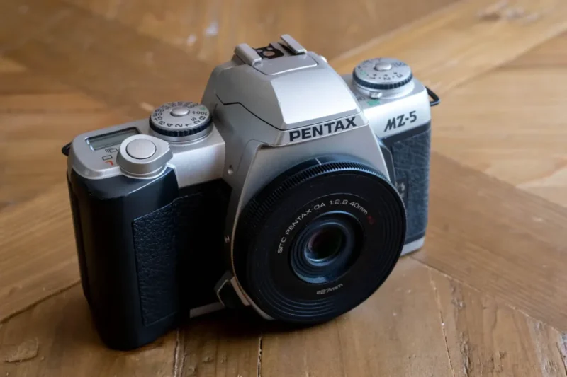 A Pentax MZ-5 film camera with a 40mm lens is placed on a wooden surface. The camera is silver and black with various dials on top.